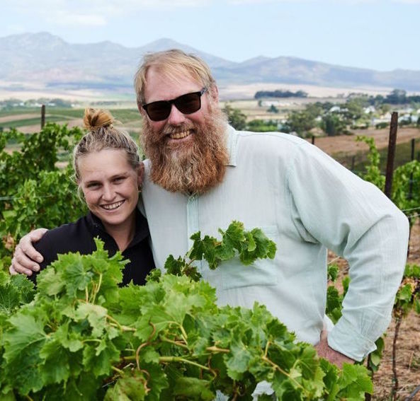 Lächelndes Winzerpaar zwischen grünen Reben, im Hintergrund hügelige Weinlandschaft unter blauem Himmel.