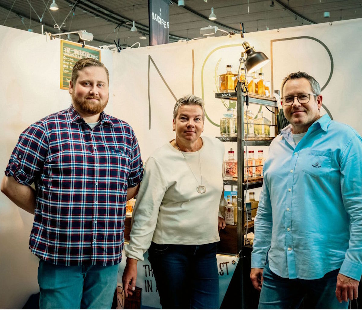 Drei Männer stehen vor einem Spirituosen-Messestand mit dem Schriftzug „NGR“ im Hintergrund, flankiert von einem beleuchteten Regal mit verschiedenen Flaschen, das auf eine Produktpräsentation im Rahmen einer Fachmesse oder Verkostung hinweist