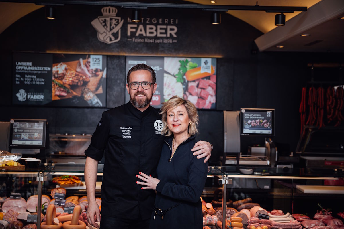 Ein Mann und eine Frau stehen lächelnd vor der Theke einer Metzgerei mit der Aufschrift „Metzgerei Faber“, im Hintergrund eine Auswahl an Fleisch- und Wurstwaren – Einblick in ein modernes Fachgeschäft für hochwertige Lebensmittel