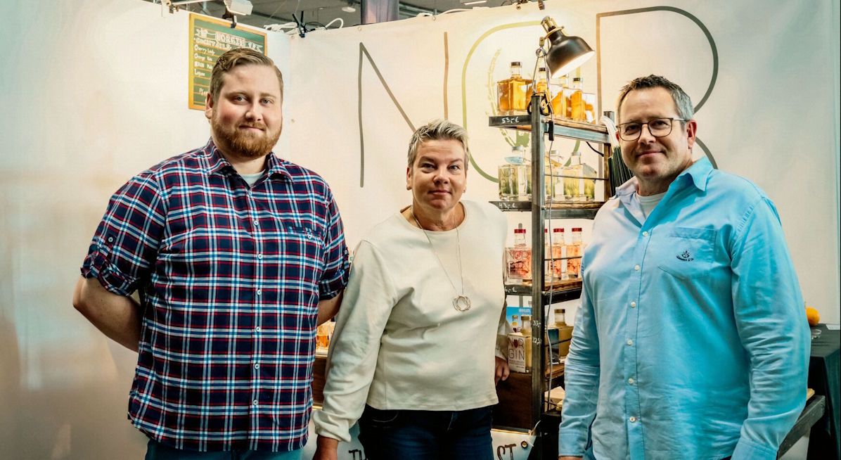 Drei Personen stehen vor einem Spirituosen-Messestand mit dem Schriftzug „NOR“ im Hintergrund, flankiert von einem beleuchteten Regal mit verschiedenen Flaschen, das auf eine Produktpräsentation im Rahmen einer Fachmesse oder Verkostung hinweist