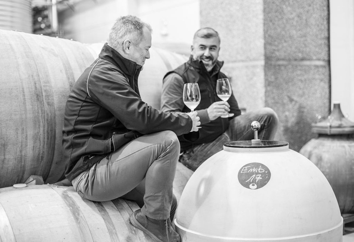Zwei Männer stoßen lachend mit Weingläsern an, sitzend auf Weinfässern in einem Weinkeller, aufgenommen in Schwarz-Weiß