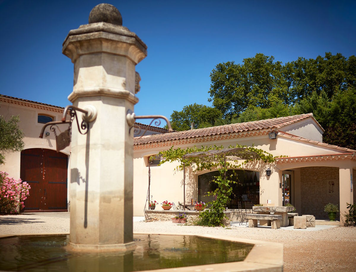 Mediterraner Weingutshof mit traditionellem Brunnen im Vordergrund und blühenden Pflanzen bei sonnigem Wetter