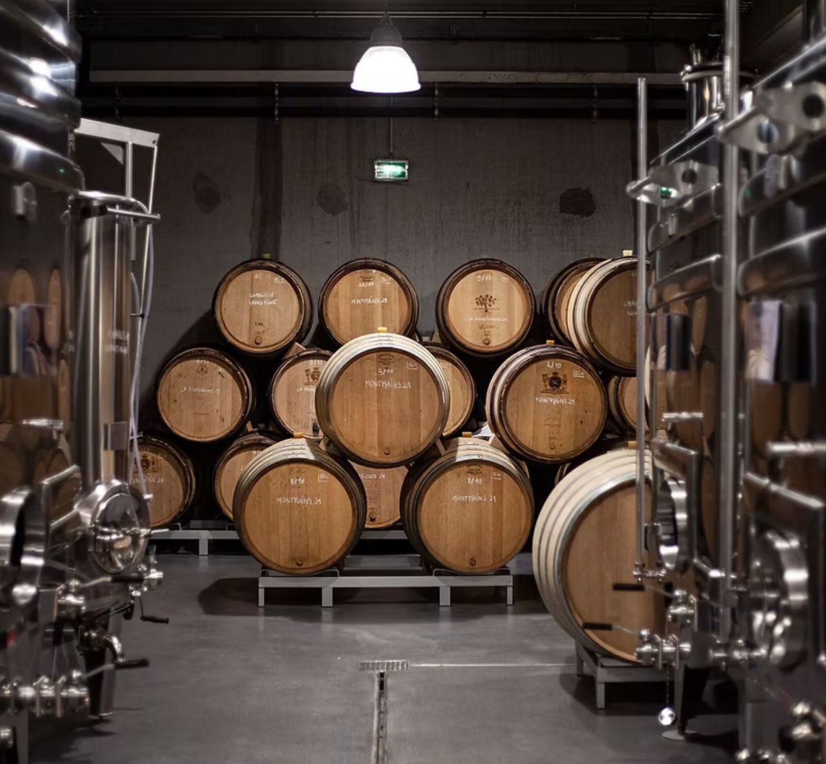 Holzfässer zur Reifung gestapelt zwischen Edelstahlbehältern in einem modernen Weinkeller.