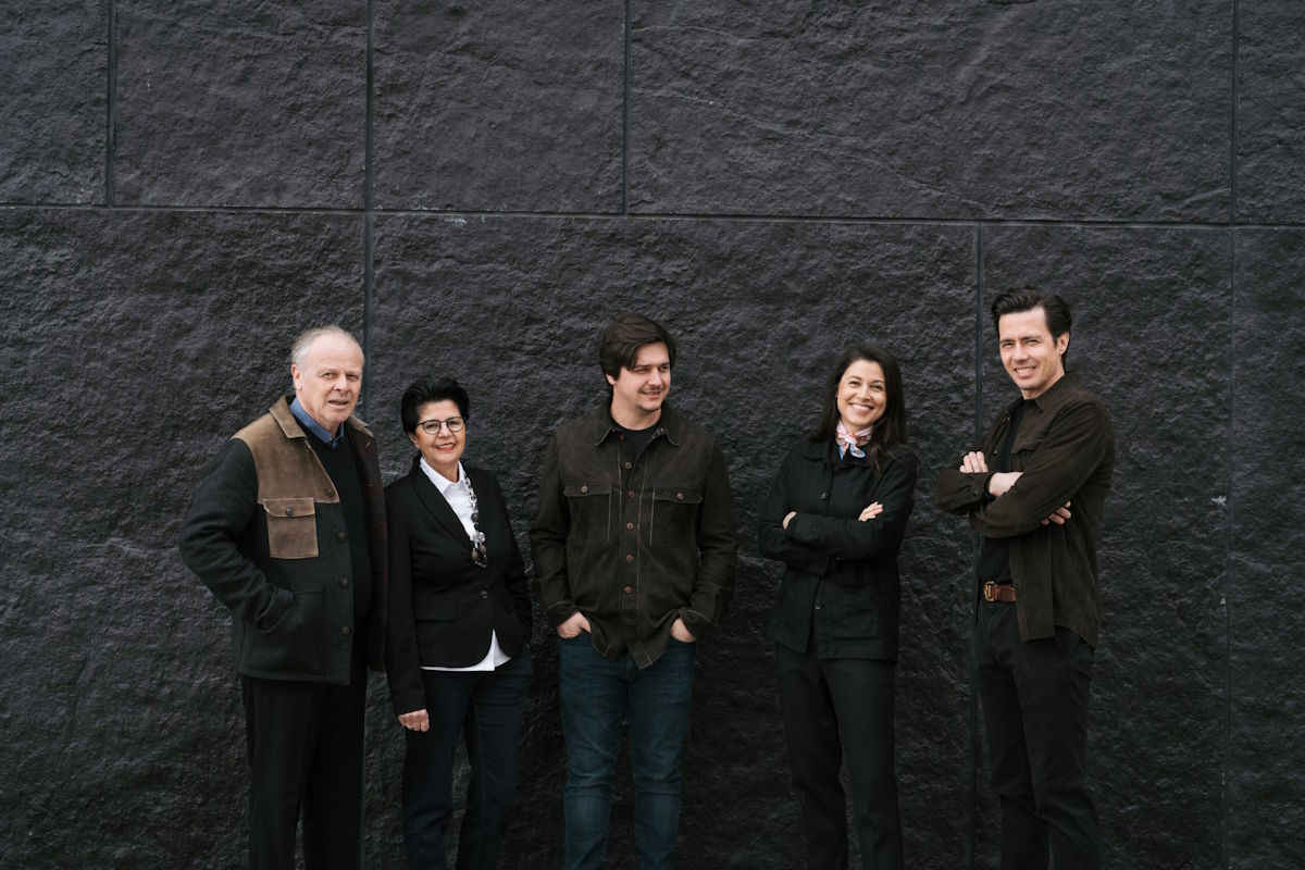  Fünf Personen in dunkler Kleidung stehen lächelnd vor einer modernen, schwarzen Steinwand