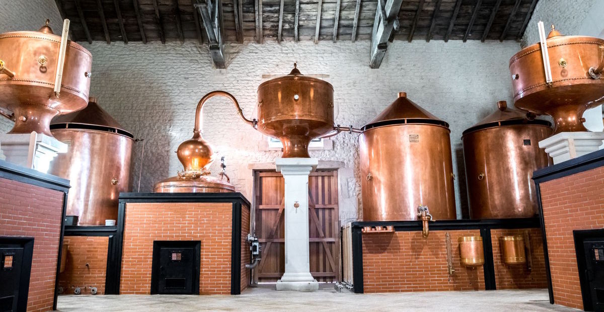 Innenansicht einer historischen Brennerei mit mehreren großen, glänzenden Kupferbrennblasen – traditionelles Handwerk in einer klassischen Destille mit gemauerten Sockeln und offenem Holzdach