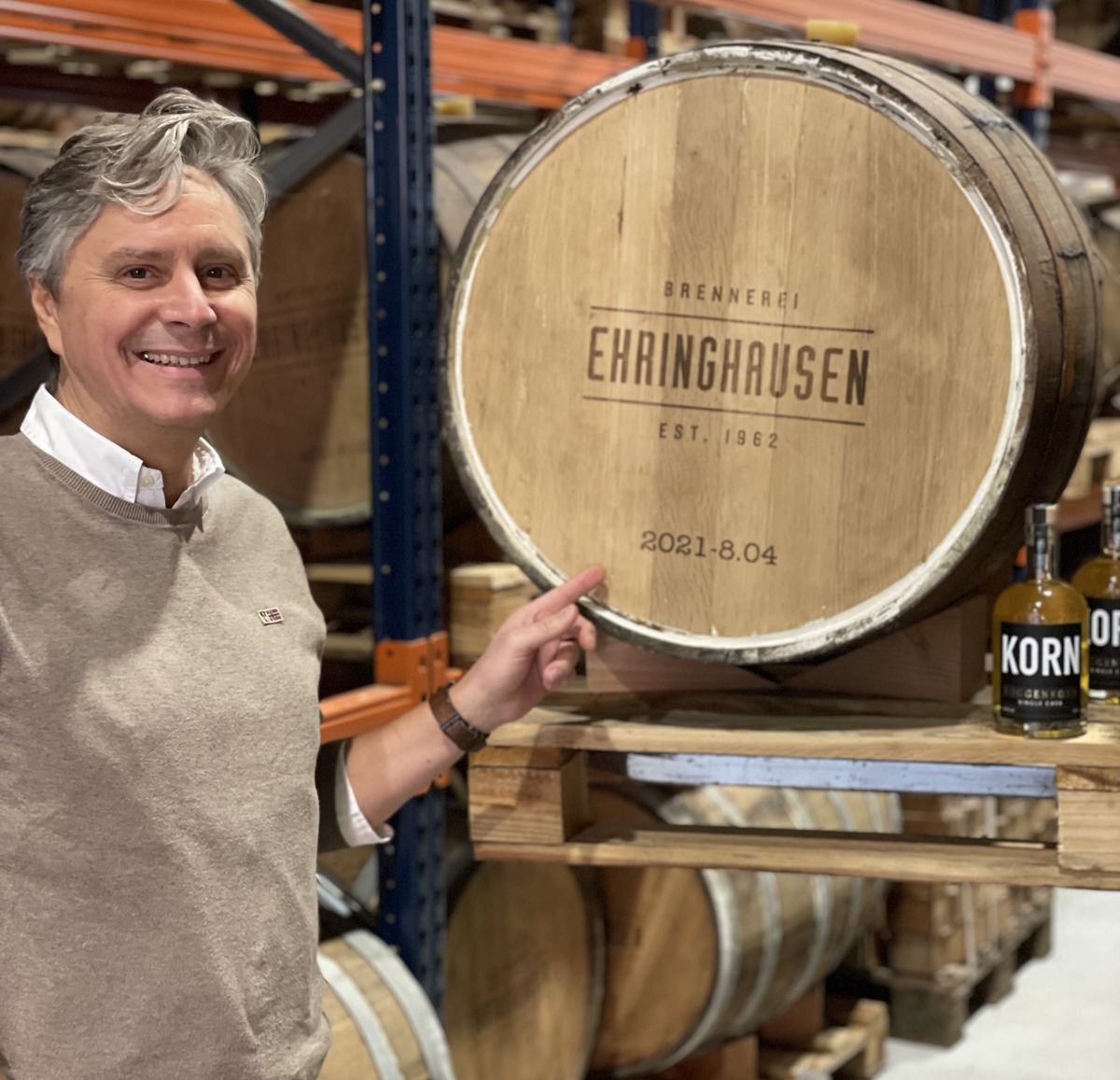 Ein Mann steht in einem Lager vor einem großen Holzfass mit dem Branding „Brennerei Ehringhausen“ und zeigt darauf; daneben stehen zwei Flaschen Kornbrand