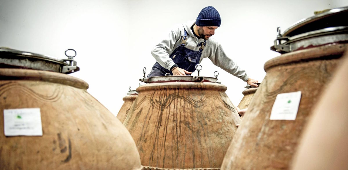 Winzer mit Mütze und Latzhose arbeitet an großen Tonamphoren mit Metallverschlüssen in einem modernen Kellerraum.