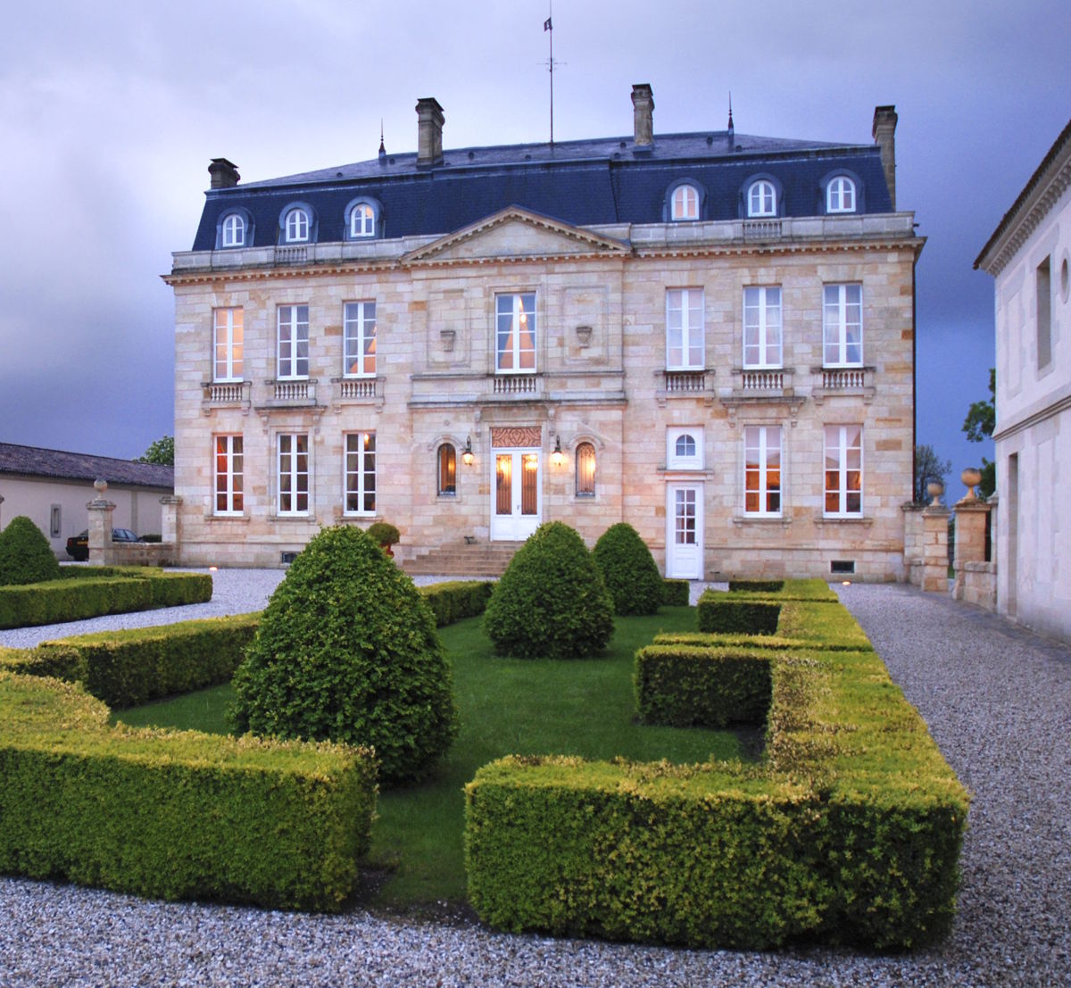 Stilvolles Château mit aufwendig gestalteter Gartenanlage aus Buchsbaumhecken und Kieswegen, vor dramatisch bewölktem Abendhimmel.