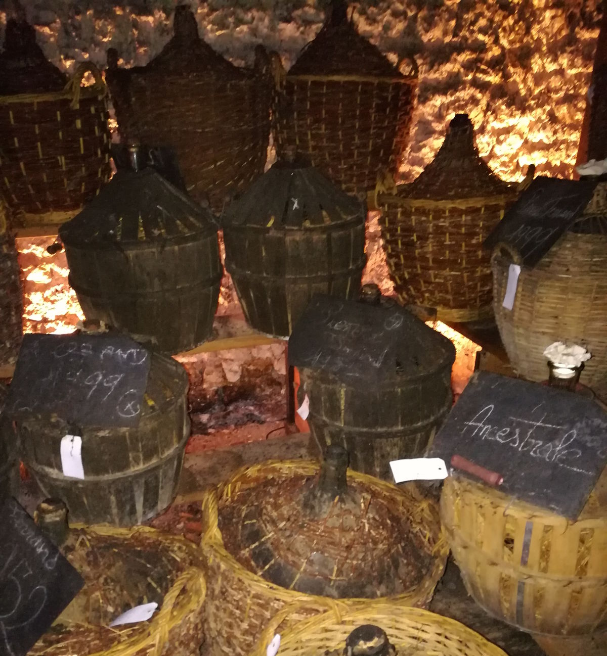 Mehrere alte Korbflaschen aus Glas mit geflochtenem Schutz in einem rustikalen Weinkeller mit Natursteinwand und stimmungsvoller Beleuchtung – einige Flaschen mit Kreidebeschriftung und Etiketten versehen