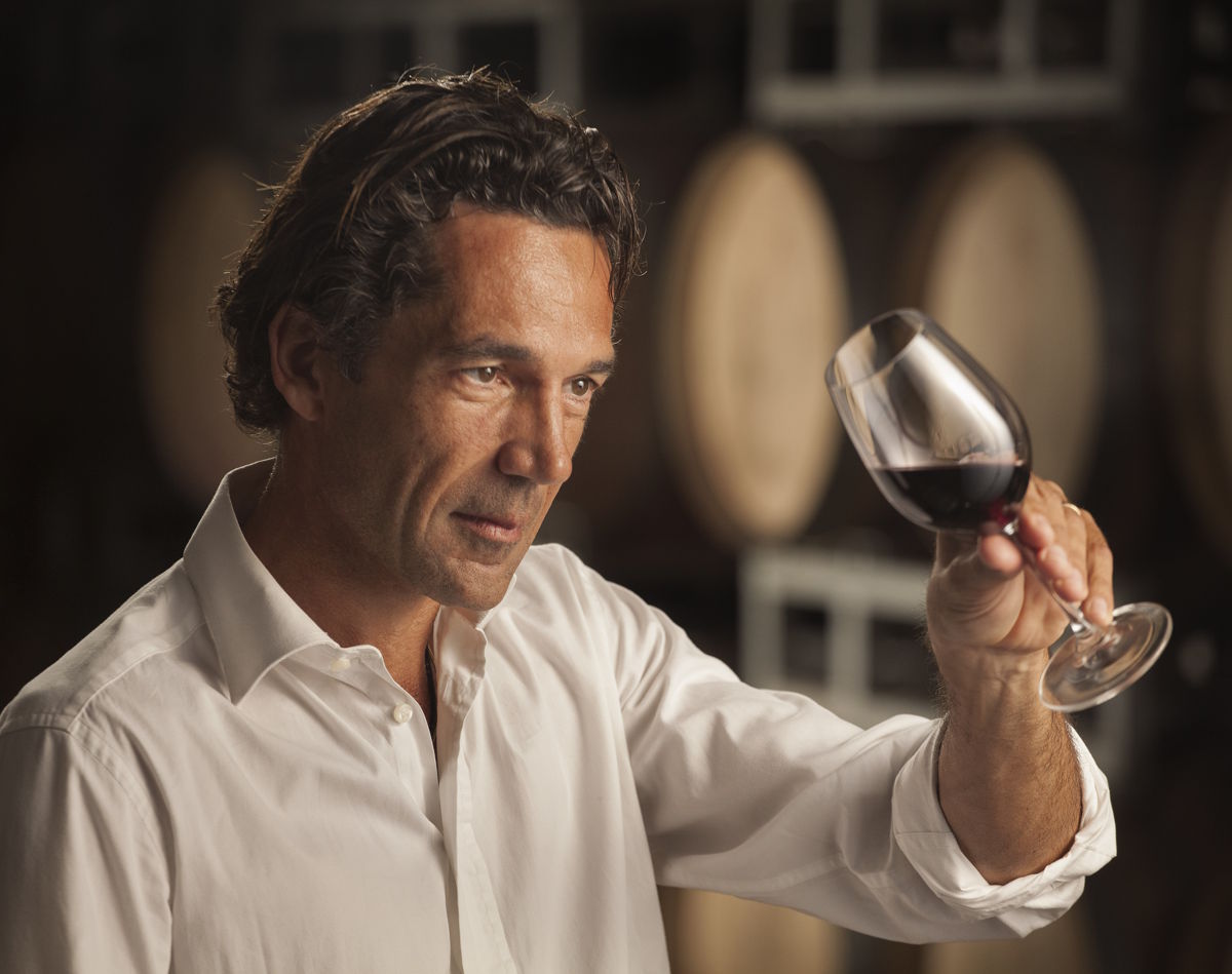 Ein Winzer in weißem Hemd betrachtet konzentriert ein Glas Rotwein vor einem Hintergrund mit Holzfässern im Weinkeller.