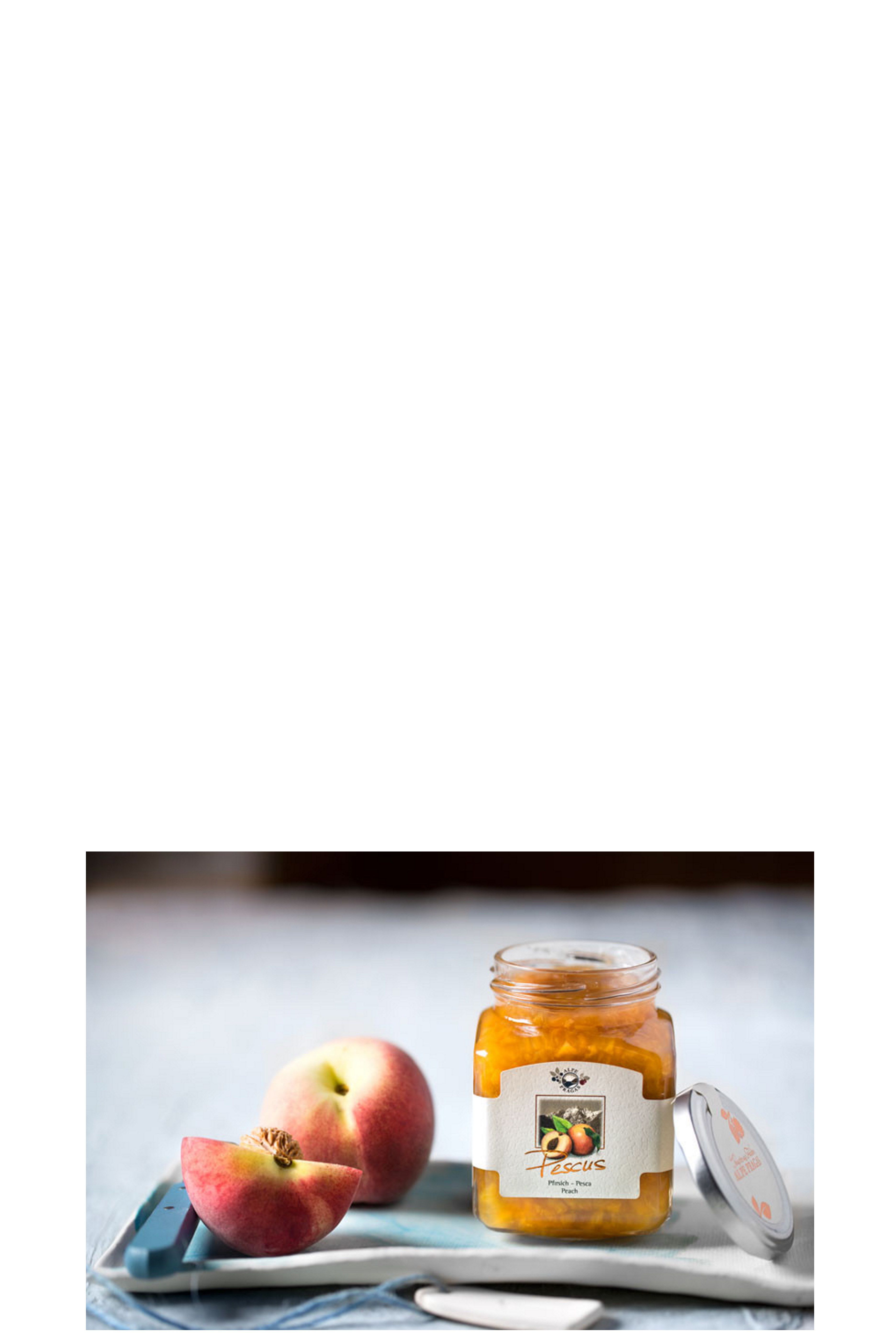 Ein sechseckiges Glas mit gold-orangefarbener Fruchtzubereitung steht geöffnet auf einem hellen Untergrund. Daneben liegt der silberne Deckel mit aprikosenfarbener Beschriftung. Im Hintergrund sind ein ganzer und ein halbierter Pfirsich zu sehen.