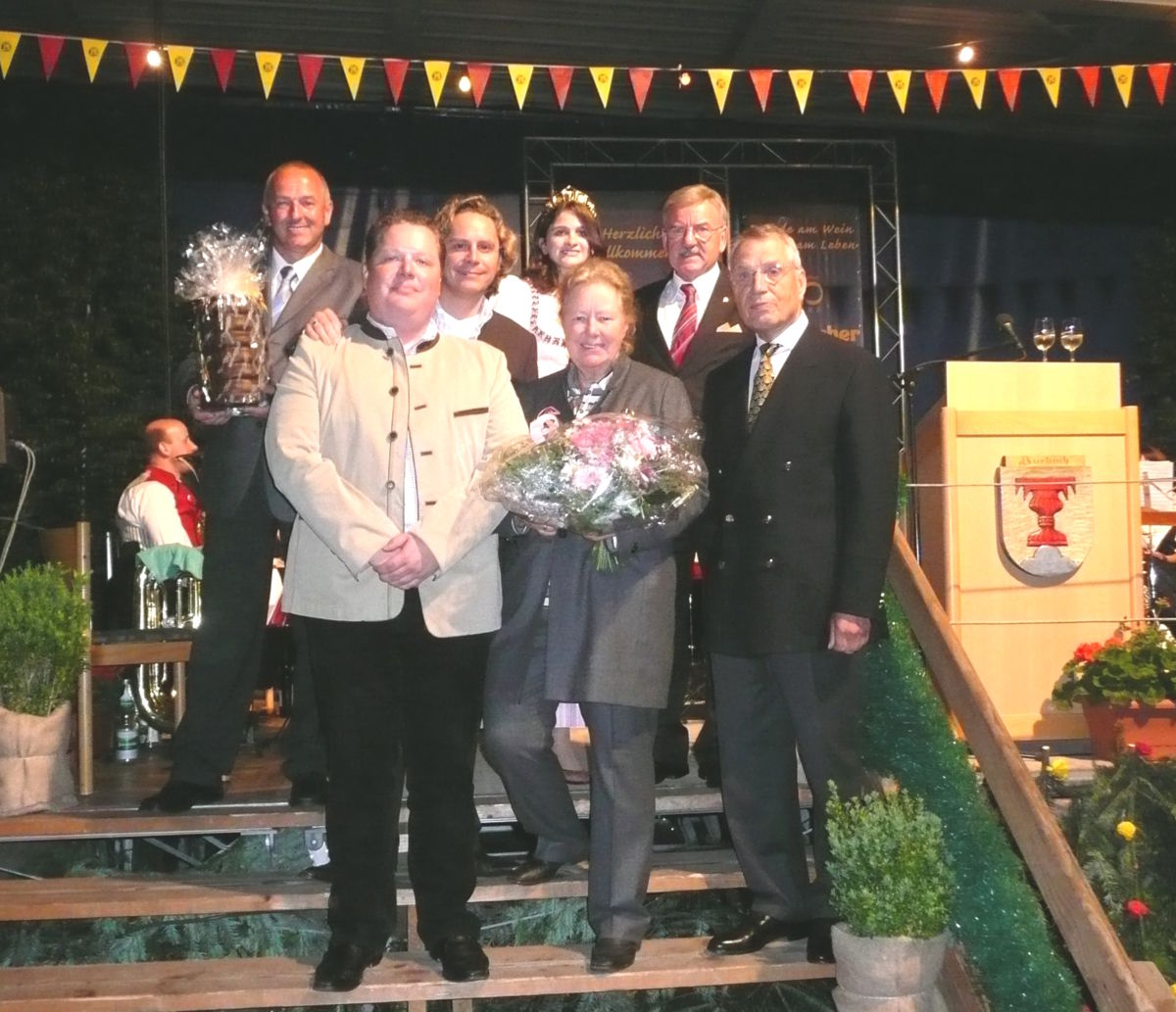  Feierliche Ehrung auf einer Bühne mit sieben Personen, darunter eine Frau mit Blumenstrauß und ein Mann mit Pokal, im Hintergrund eine Weinprinzessin und ein Rednerpult