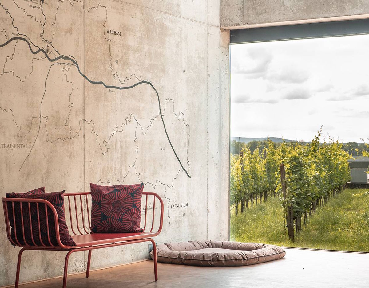 Moderner Innenraum mit roter Sitzbank und Wandkarte, daneben ein großes offenes Fenster mit Blick auf einen angrenzenden Weinberg.