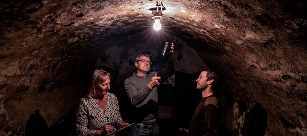 Drei Personen in einem alten, gewölbten Weinkeller, eine Flasche wird gegen das Licht gehalten und begutachtet.