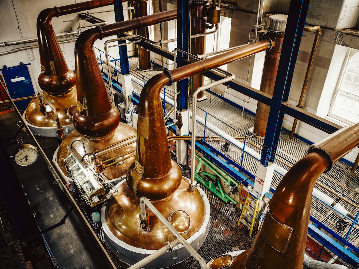 Innenansicht einer Whiskybrennerei mit mehreren glänzenden Kupferbrennblasen in einer industriellen Produktionshalle – klassische Pot Stills für die Destillation von Whisky, umgeben von Rohren, Plattformen und Maschinen in typischer Destillerie-Architektu