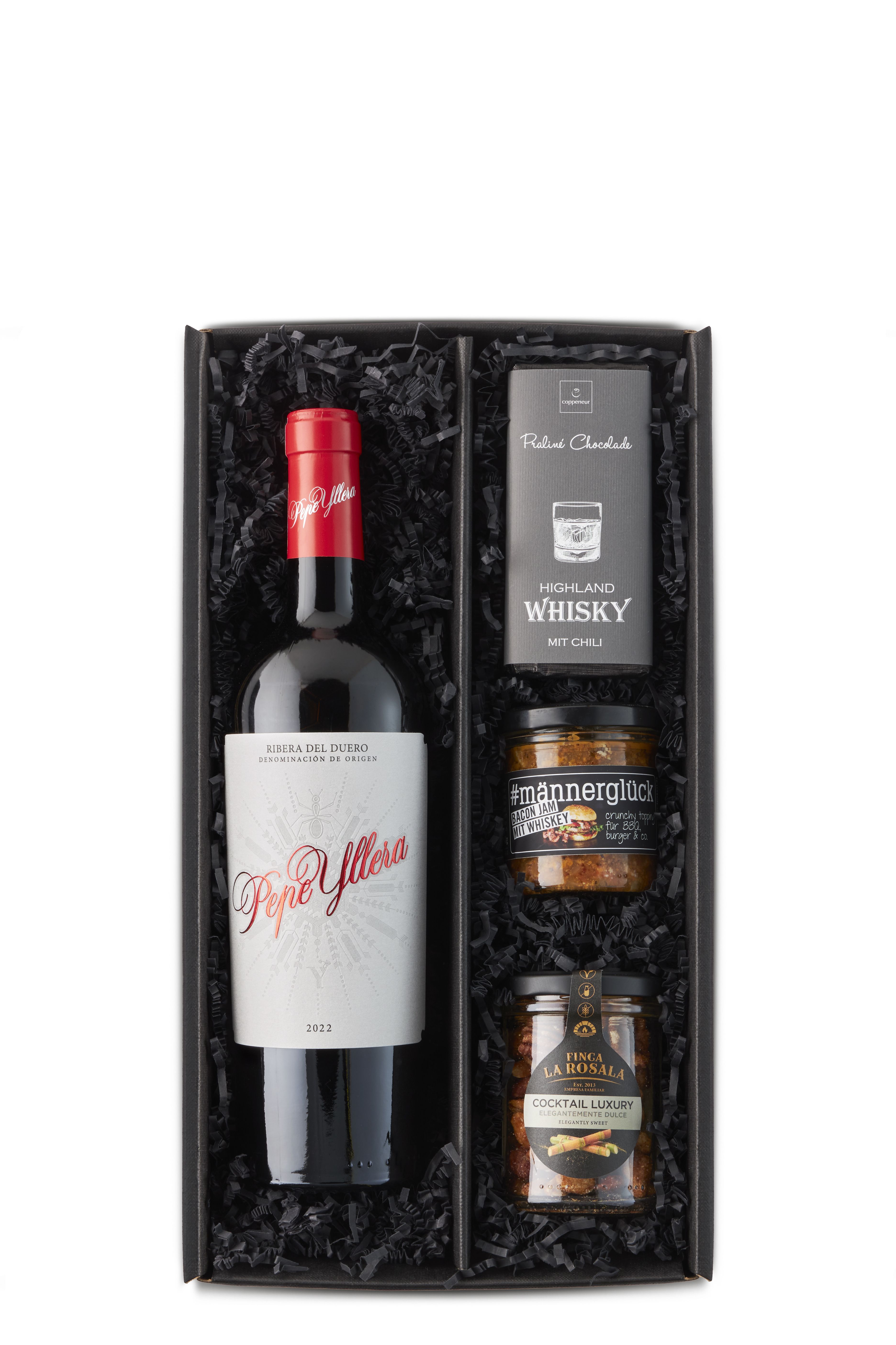 Diese schwarze Geschenkbox enthält eine Flasche Rotwein „Pepe Yllera“, eine Tafel Highland Whisky-Schokolade mit Chili, ein Glas Bacon Jam mit Whiskey („#männerglück“) sowie einen Luxus-Nusscocktail – gebettet in schwarzem Füllmaterial