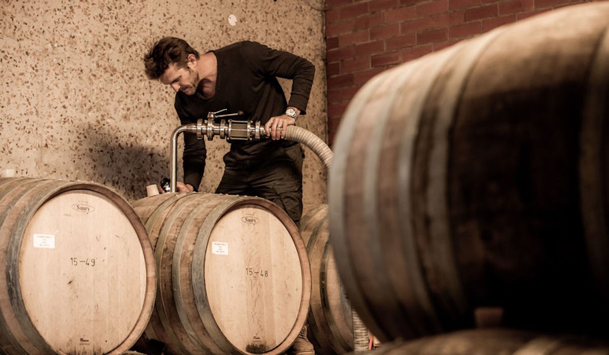 Winzer bei der Arbeit im Weinkeller, füllt Wein mithilfe eines Schlauchs in Holzfässer – umgeben von mehreren Eichenfässern.