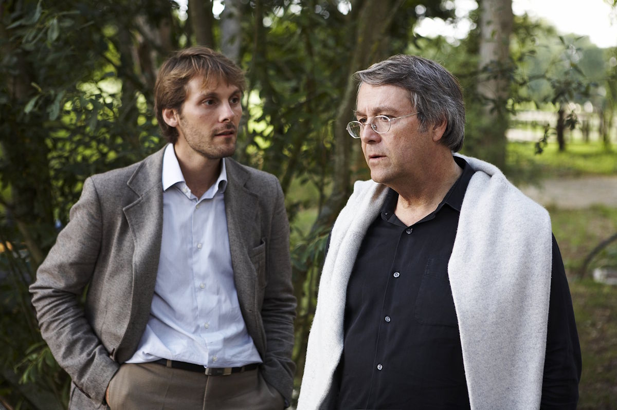 Zwei Männer, einer jüngeren und einer älteren Generation, stehen in einem Garten oder Park und führen ein Gespräch – beide elegant gekleidet, mit Bäumen und Natur im Hintergrund