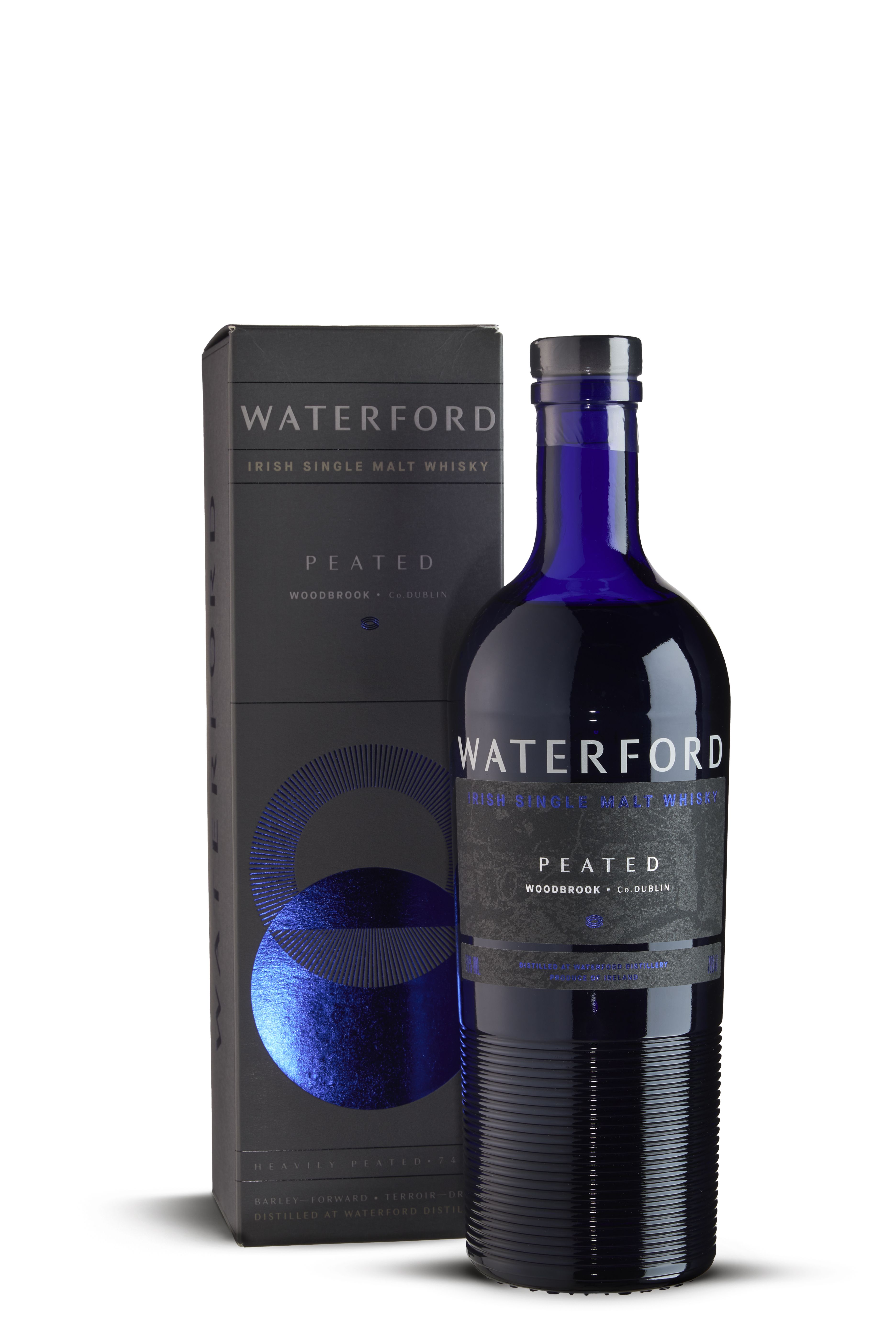 Eine dunkle, glänzende Flasche aus tiefblauem Glas mit silber-weißer Aufschrift „WATERFORD“, daneben eine schwarze Verpackung mit silbernem und blauem Aufdruck, darunter Kreis-Motive und Text. Flasche hat gerippte Struktur im unteren Teil.