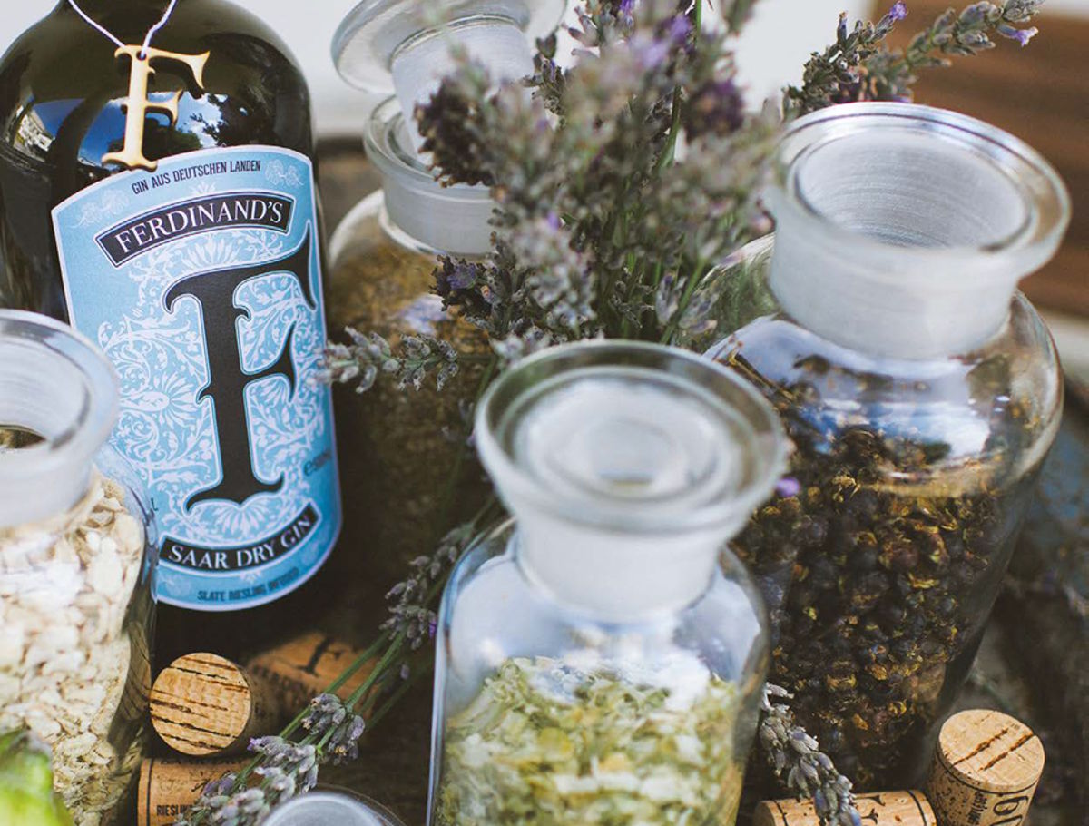 Nahaufnahme einer Flasche Ferdinand’s Saar Dry Gin umgeben von verschiedenen getrockneten Botanicals wie Lavendel, Hopfen und Kräutern in Glasgefäßen auf einem rustikalen Untergrund
