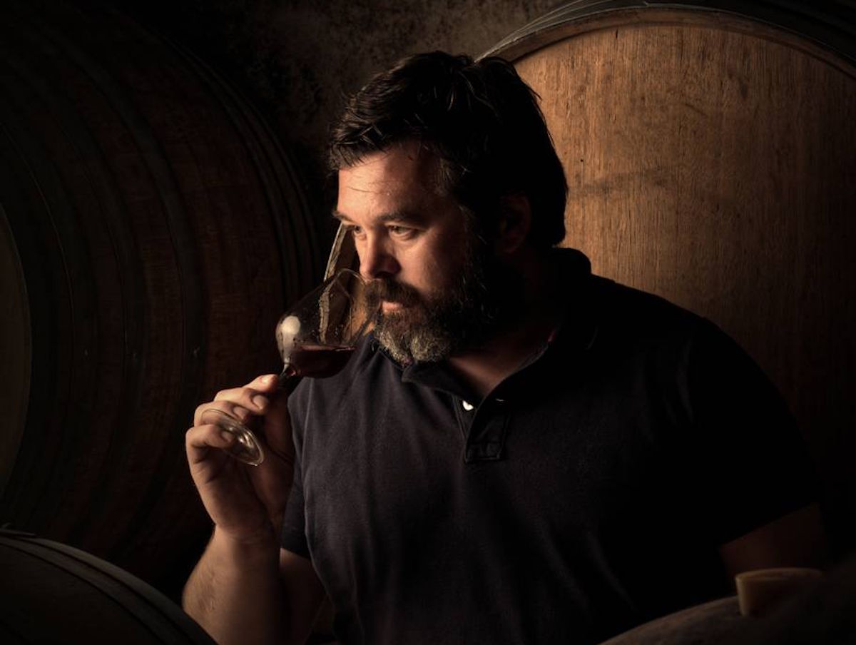  Mann mit Bart riecht konzentriert an einem Glas Rotwein vor großen Holzfässern in stimmungsvoller Kelleratmosphäre