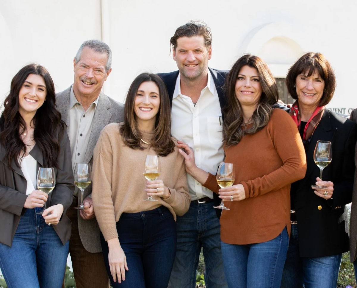 Siebenköpfige Winzerfamilie steht lachend zusammen und hält Weißweingläser in der Hand
