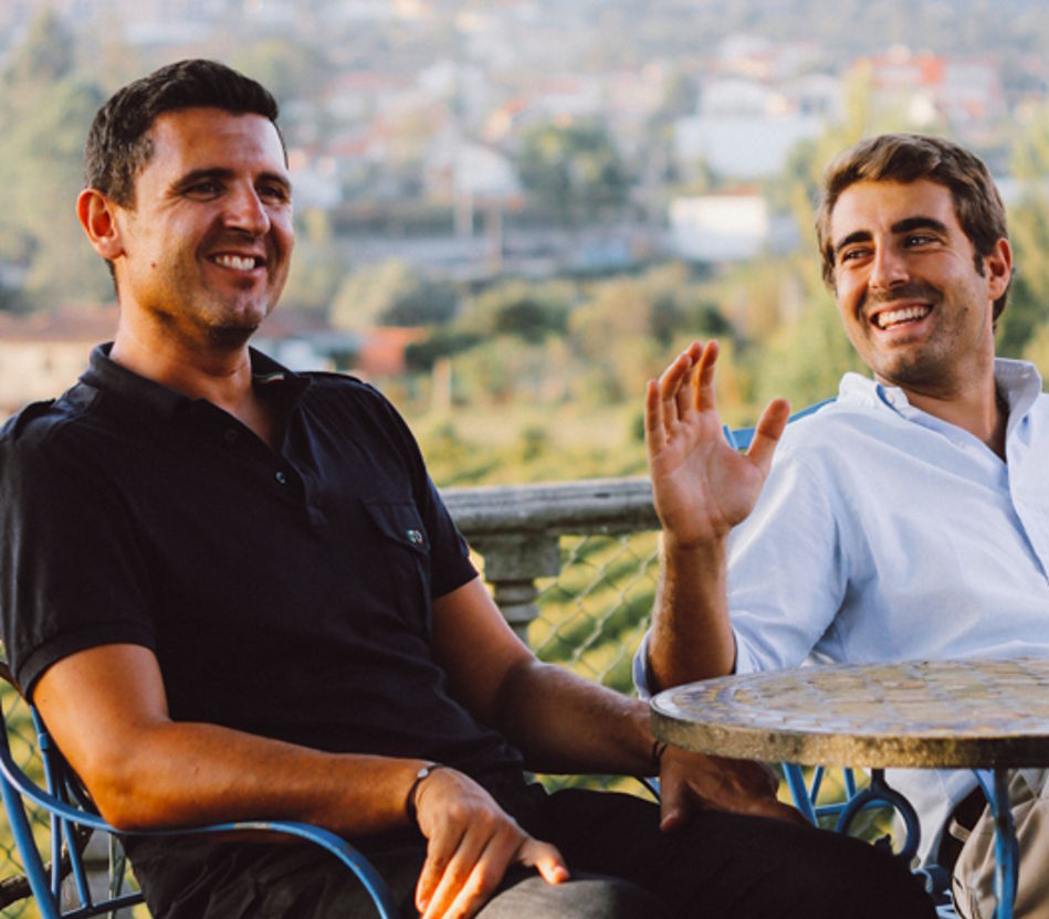 Zwei Winzer sitzen lachend an einem Gartentisch mit Blick auf die Weinberge im Hintergrund.