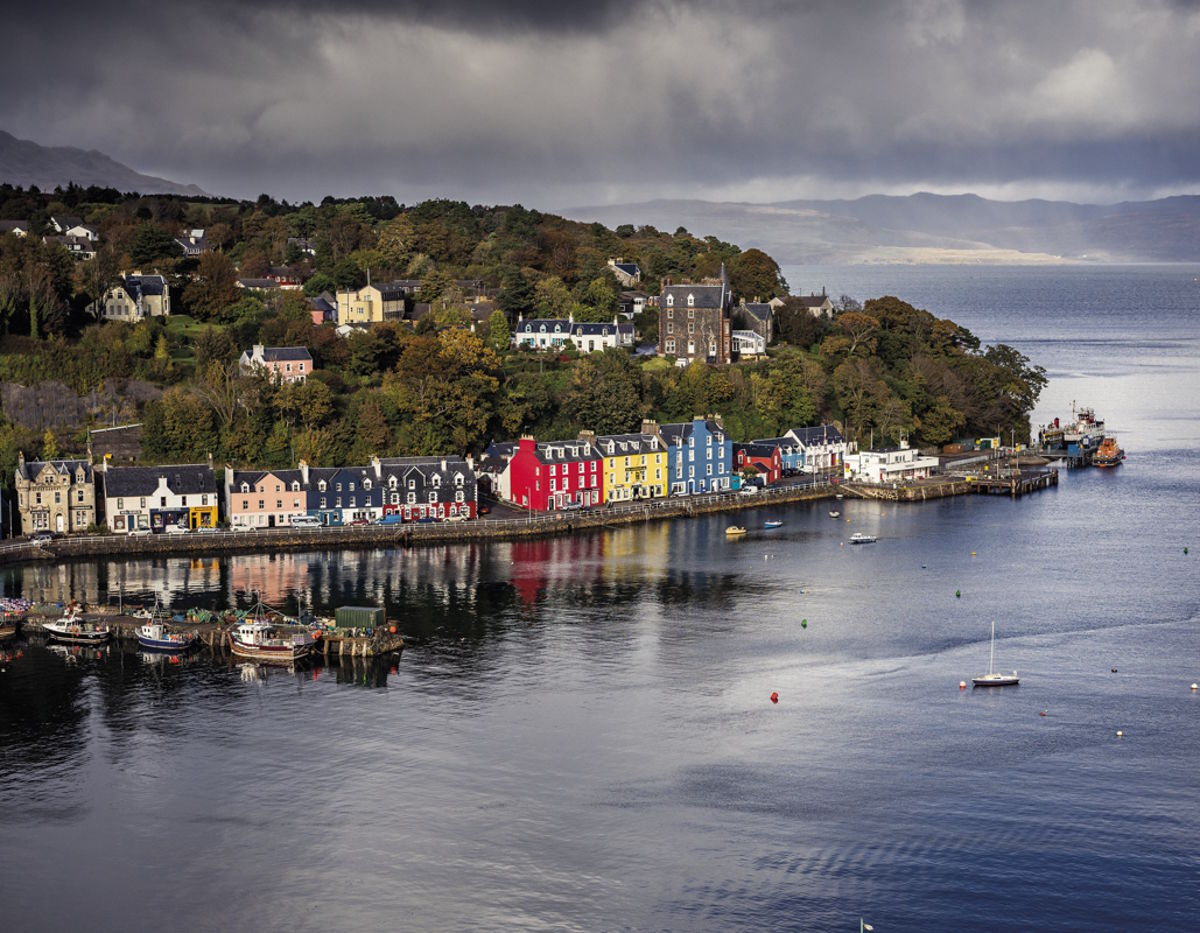 Blick auf den malerischen Hafen von Tobermory auf der Isle of Mull in Schottland mit seinen bunten Häuserfassaden entlang der Küste, ruhigem Wasser im Vordergrund und ein bewölkter Himmel im Hintergrund