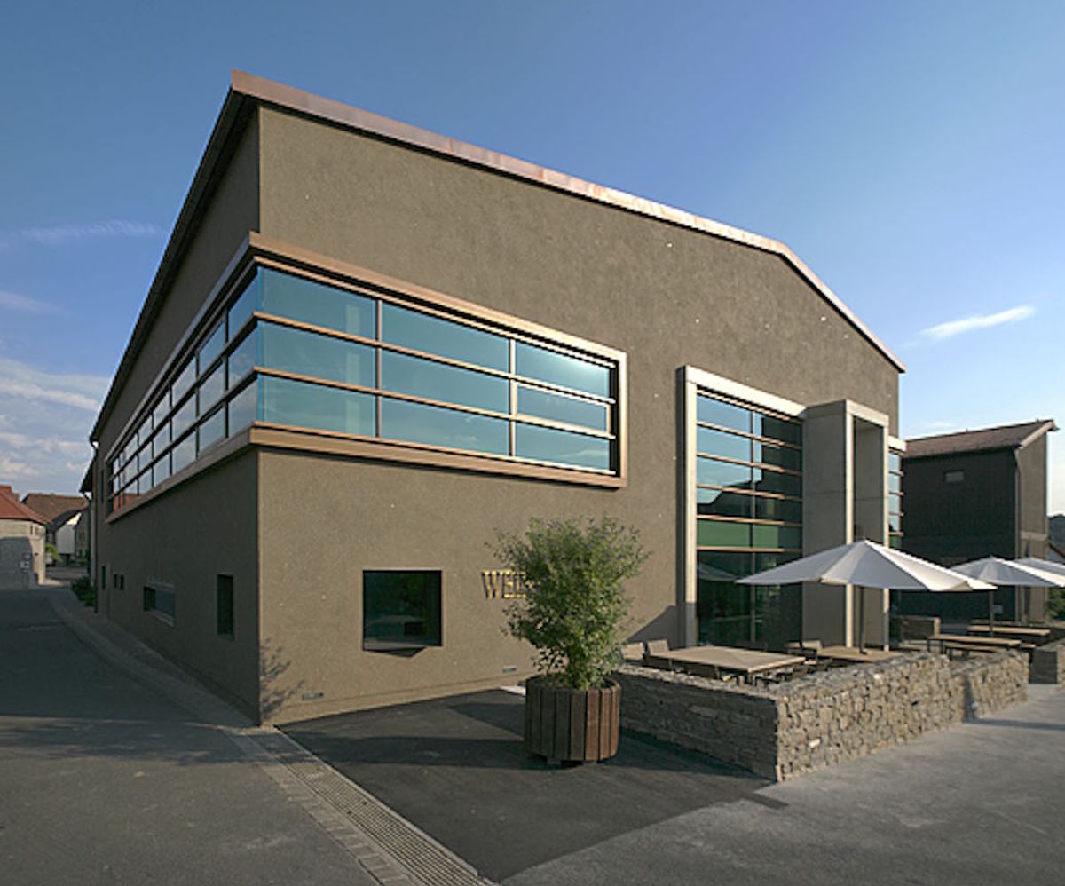  Modernes Weingutsgebäude mit großen Glasfronten, klarer Architektur und Terrasse mit Sonnenschirmen