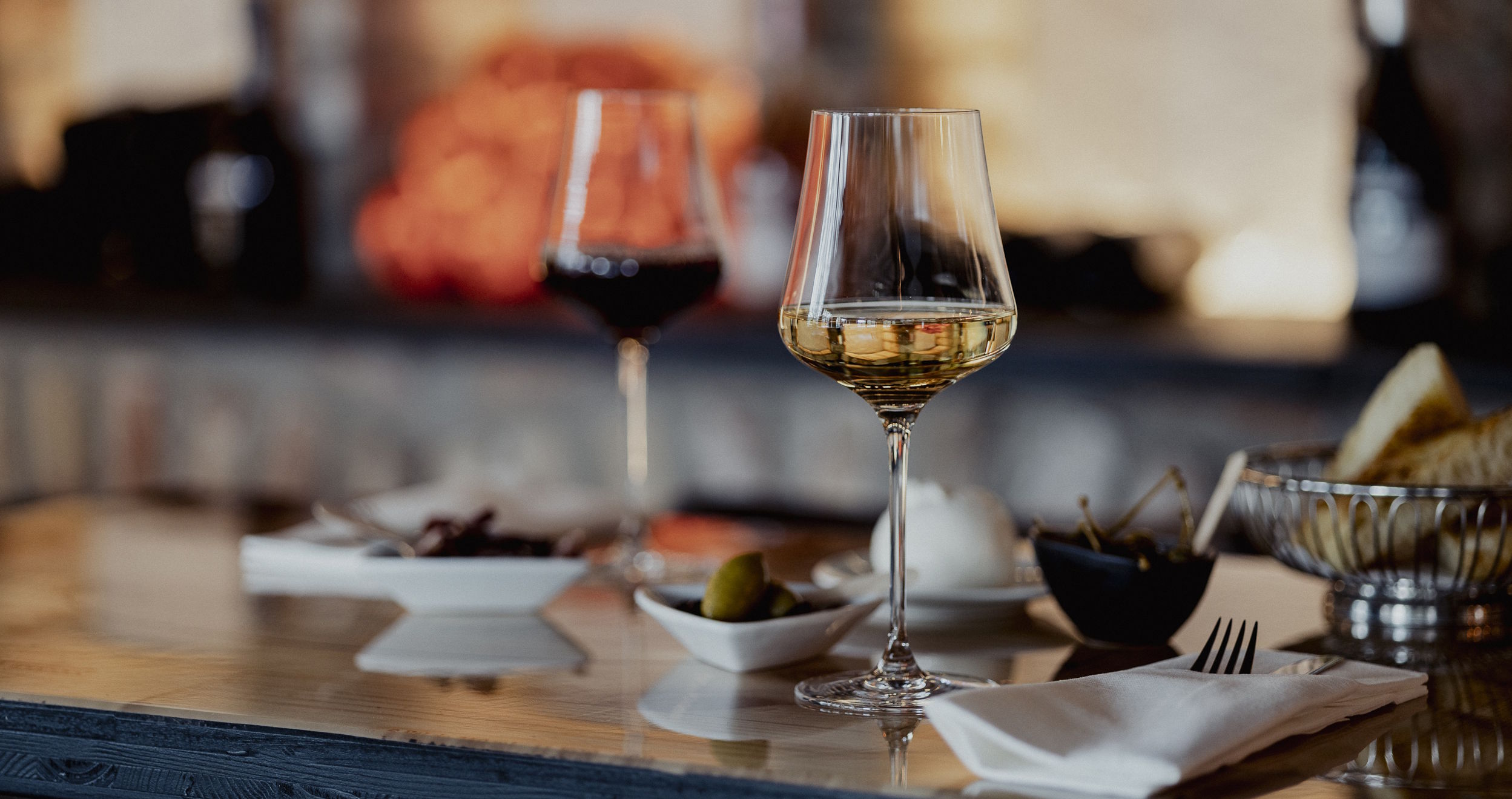 Ein Glas Weißwein und ein Glas Rotwein stehen auf einem gedeckten Tisch mit kleinen Speisen und Beilagen – stilvolle Verkostungsszene mit Fokus auf Genuss, Foodpairing und hochwertige Weine
