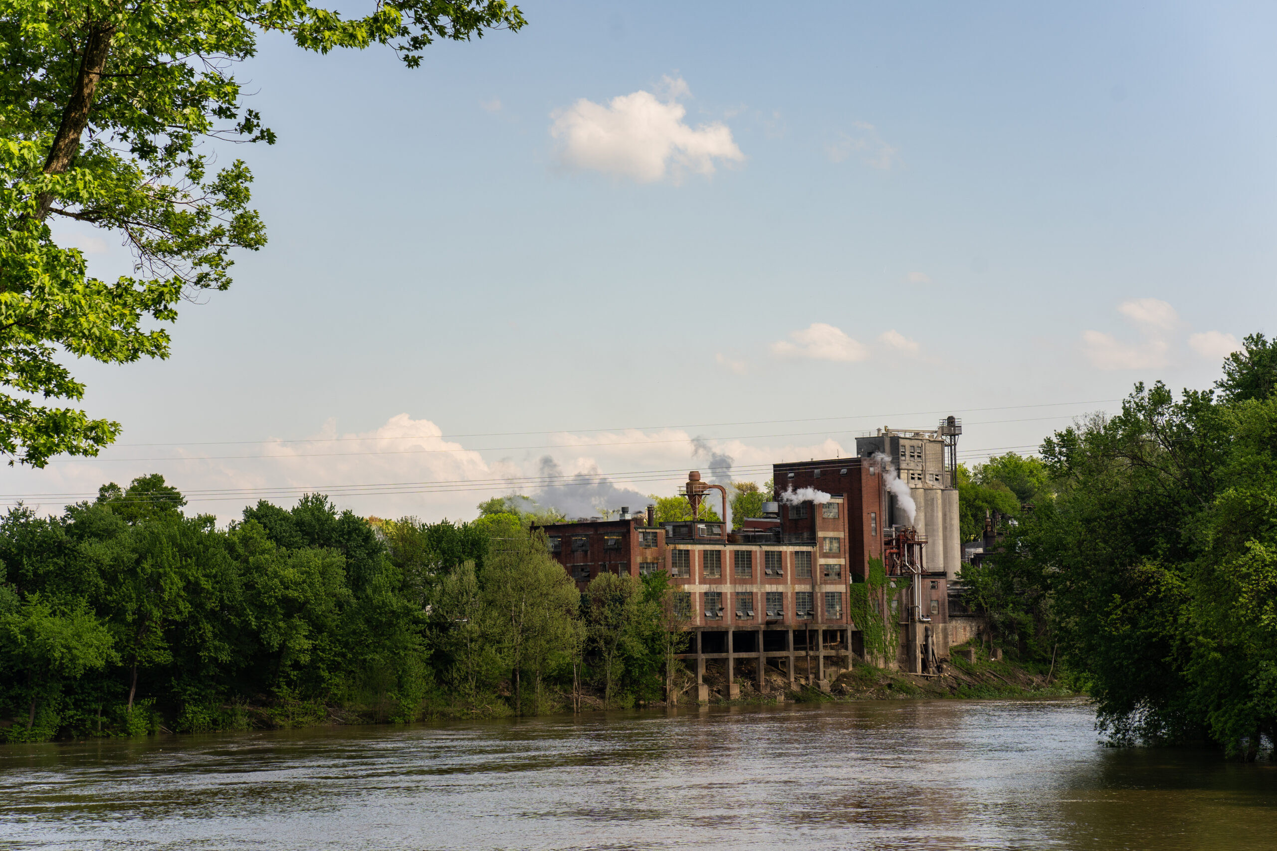 Ein größeres Industriegebäude steht direkt an einem Fluss und ist von dichter, grüner Vegetation umgeben – im Vordergrund ruhiges Wasser, im Hintergrund eine Mischung aus Natur und industrieller Architektur