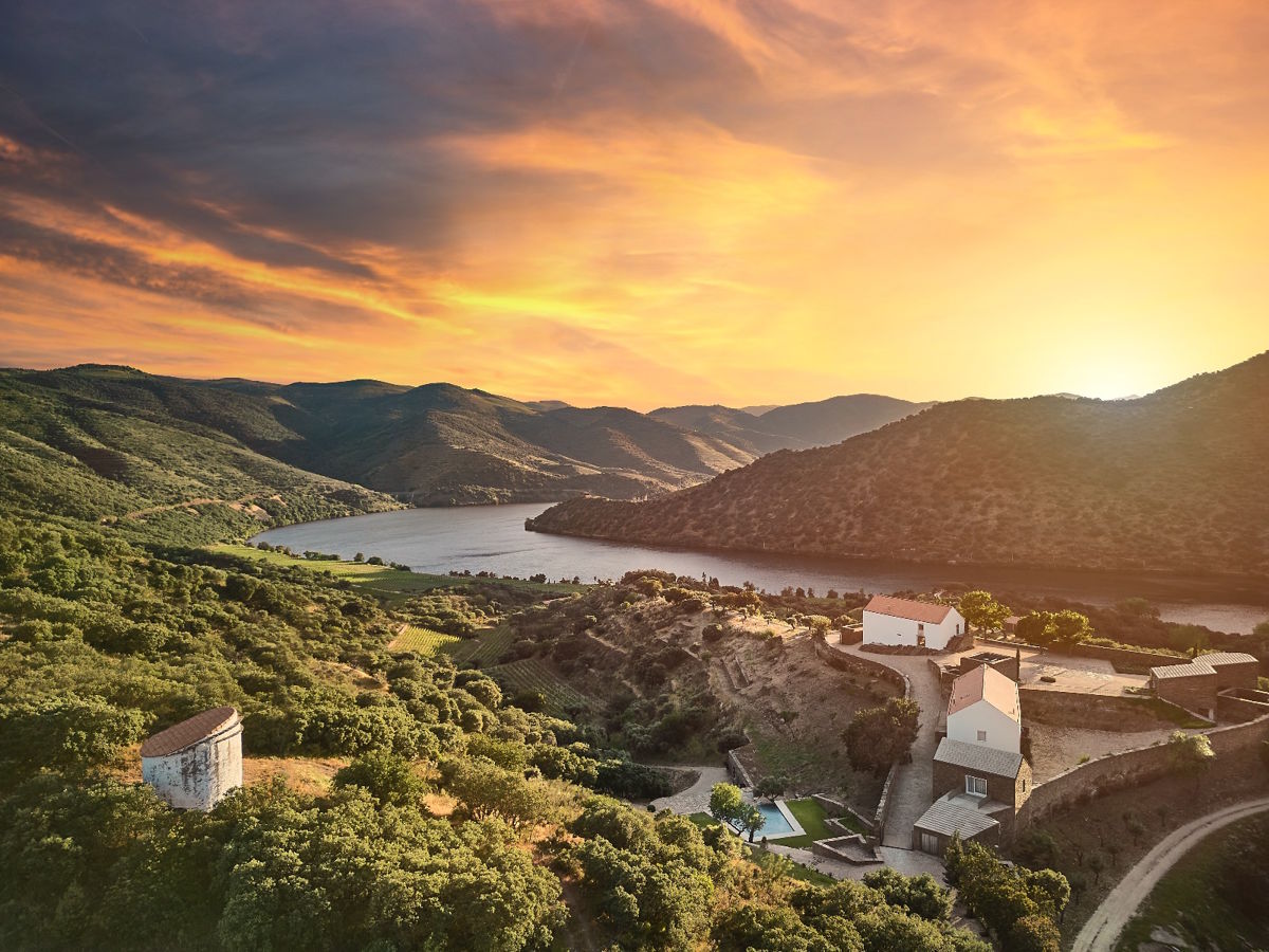 Luftaufnahme einer hügeligen Flusslandschaft bei Sonnenuntergang mit verstreuten Gebäuden und dichter Vegetation im Vordergrund.