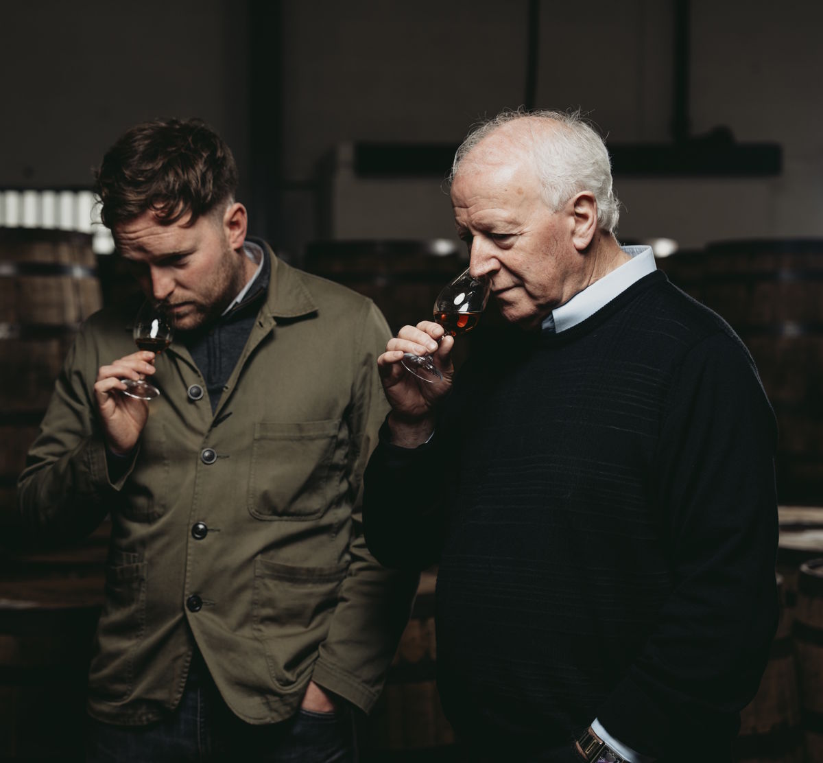Zwei Männer in einem Fasslager verkosten konzentriert einen dunklen Whisky – sie halten ein Glas an die Nase und bewerten den Duft, um die Aromenvielfalt und Qualität des Destillats zu beurteilen