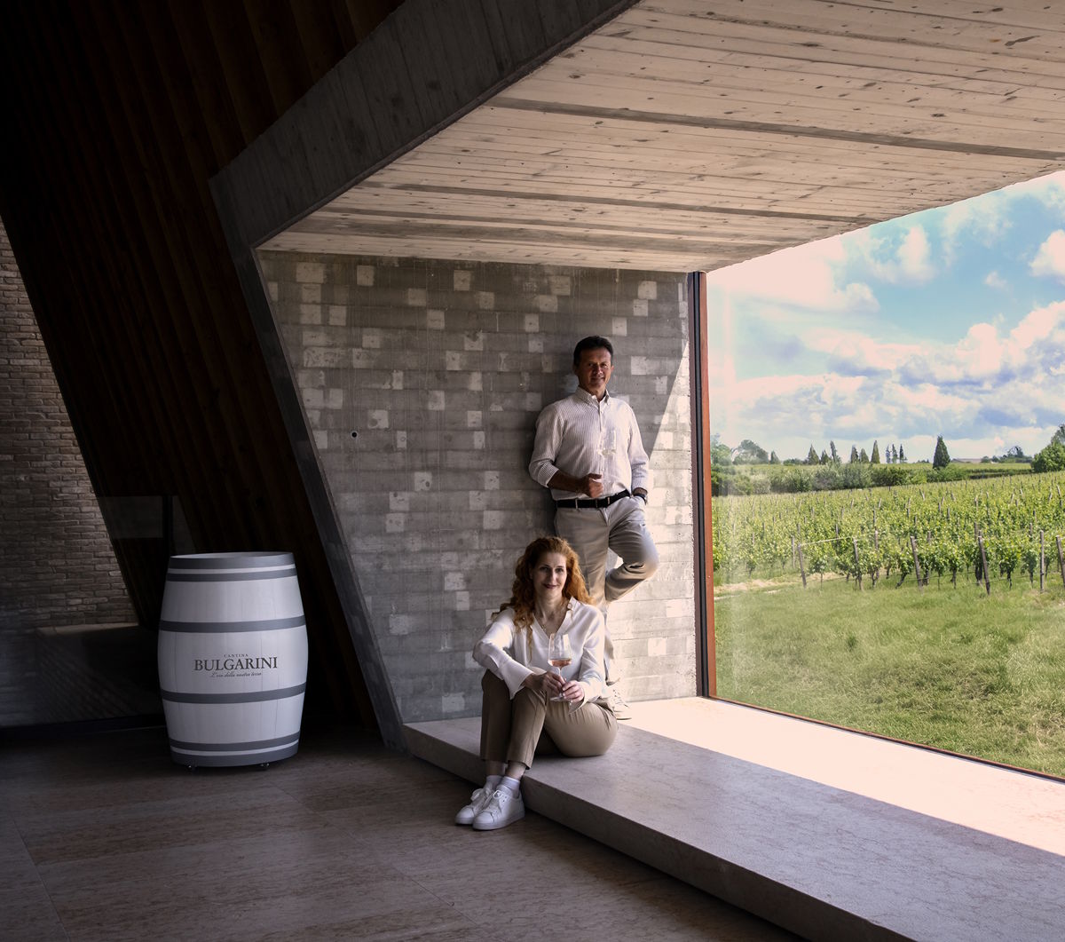 Architektur trifft Weinbau – Ausblick vom modernen Weingut in die Reben Zwei Personen in modernem Innenraum mit großem Fenster und Blick auf einen Weinberg, daneben ein Fass mit Aufschrift „Bulgari“.