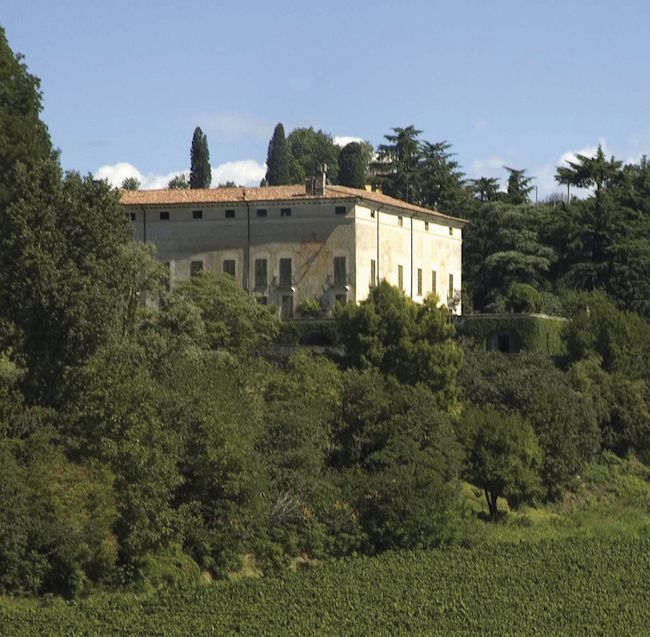 Historisches Gutshaus mit rotem Ziegeldach auf einem Hügel, umgeben von Weinbergen und Zypressen.