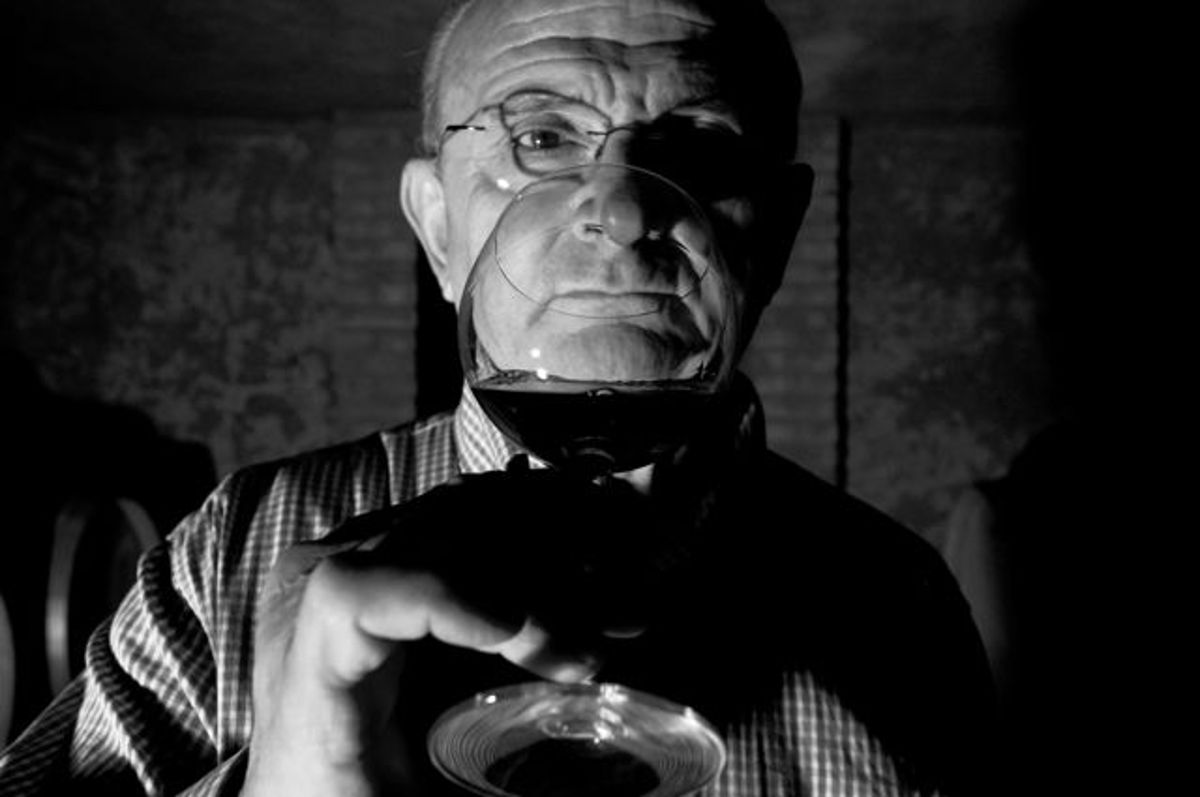 Älterer Mann mit Brille betrachtet konzentriert ein Glas Rotwein im Halbdunkel eines Weinkellers, stimmungsvolle Schwarz-Weiß-Aufnahme