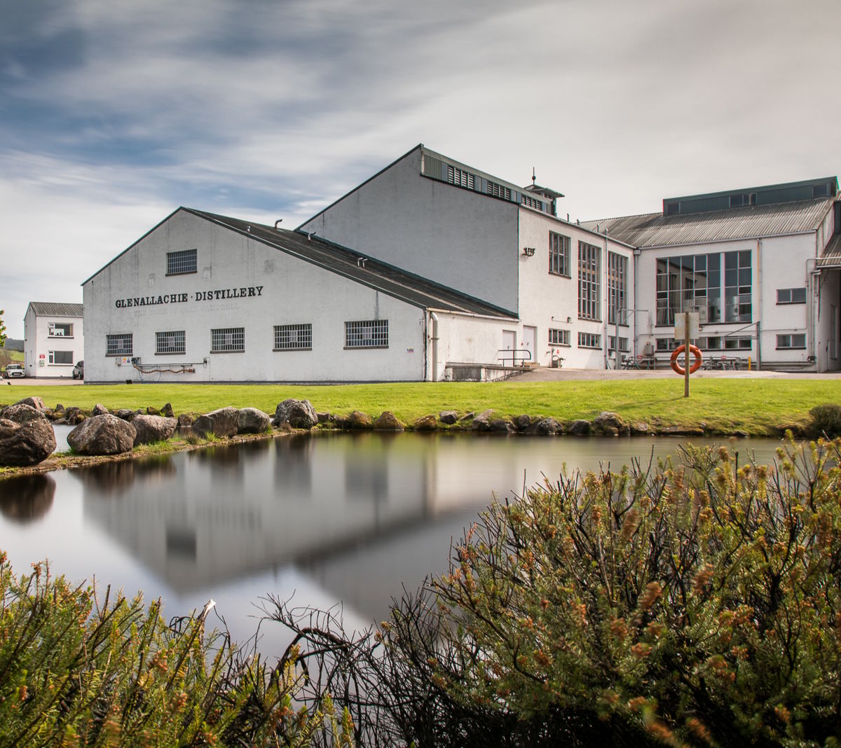 Blick auf das Gebäude der GlenAllachie Distillery inmitten der schottischen Natur, mit gepflegtem Rasen, einem kleinen Teich im Vordergrund und bewölktem Himmel – eine traditionsreiche Whiskybrennerei in der Region Speyside
