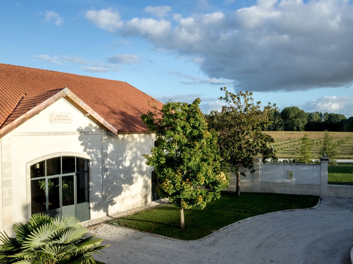 Teilansicht eines hellen Gebäudes mit rotem Ziegeldach, umgeben von gepflegtem Grün, Bäumen und einer weißen Mauer – im Hintergrund Felder und ein weitläufiger Himmel mit Wolken