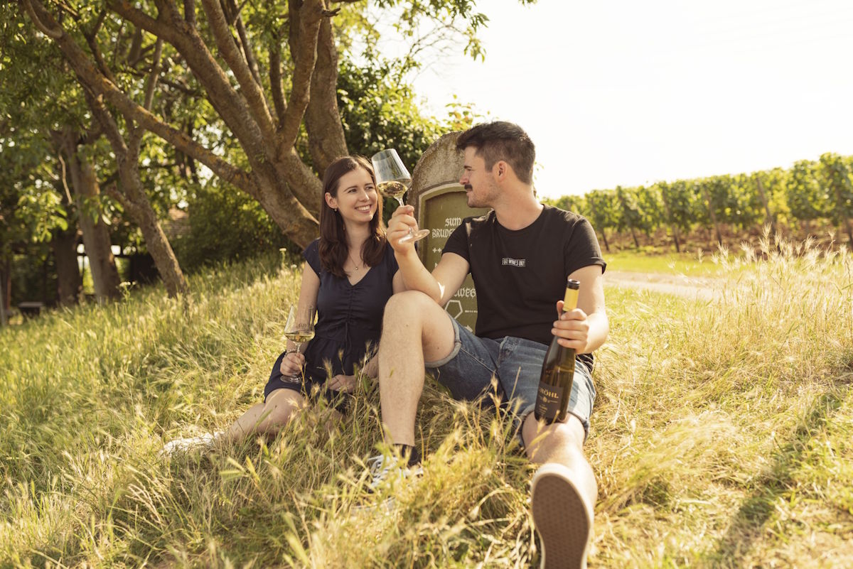 Junges Paar sitzt entspannt mit einem Glas Wein auf einer Wiese am Weinberg und stößt an, umgeben von sommerlicher Natur