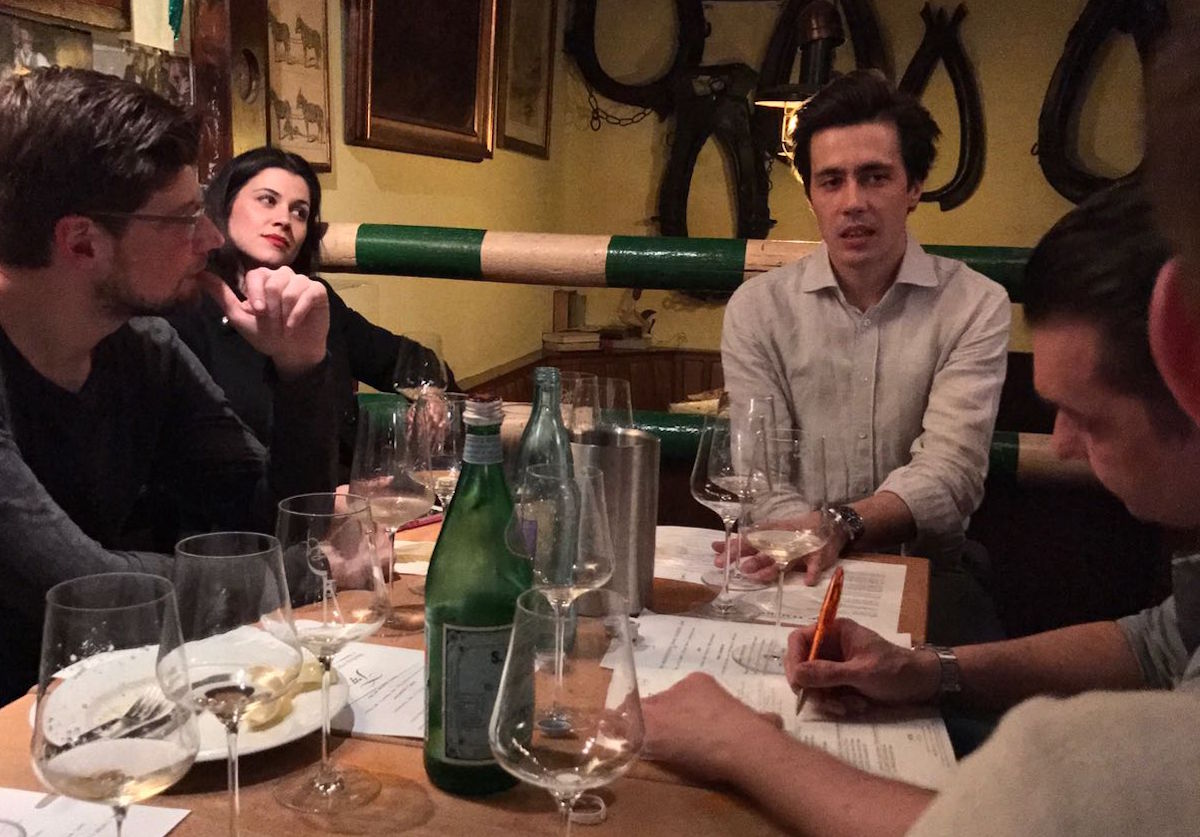  Fünf Personen sitzen bei einer Weinprobe am Tisch mit gefüllten Gläsern, Wasserflaschen und Verkostungsnotizen in gemütlichem Ambiente