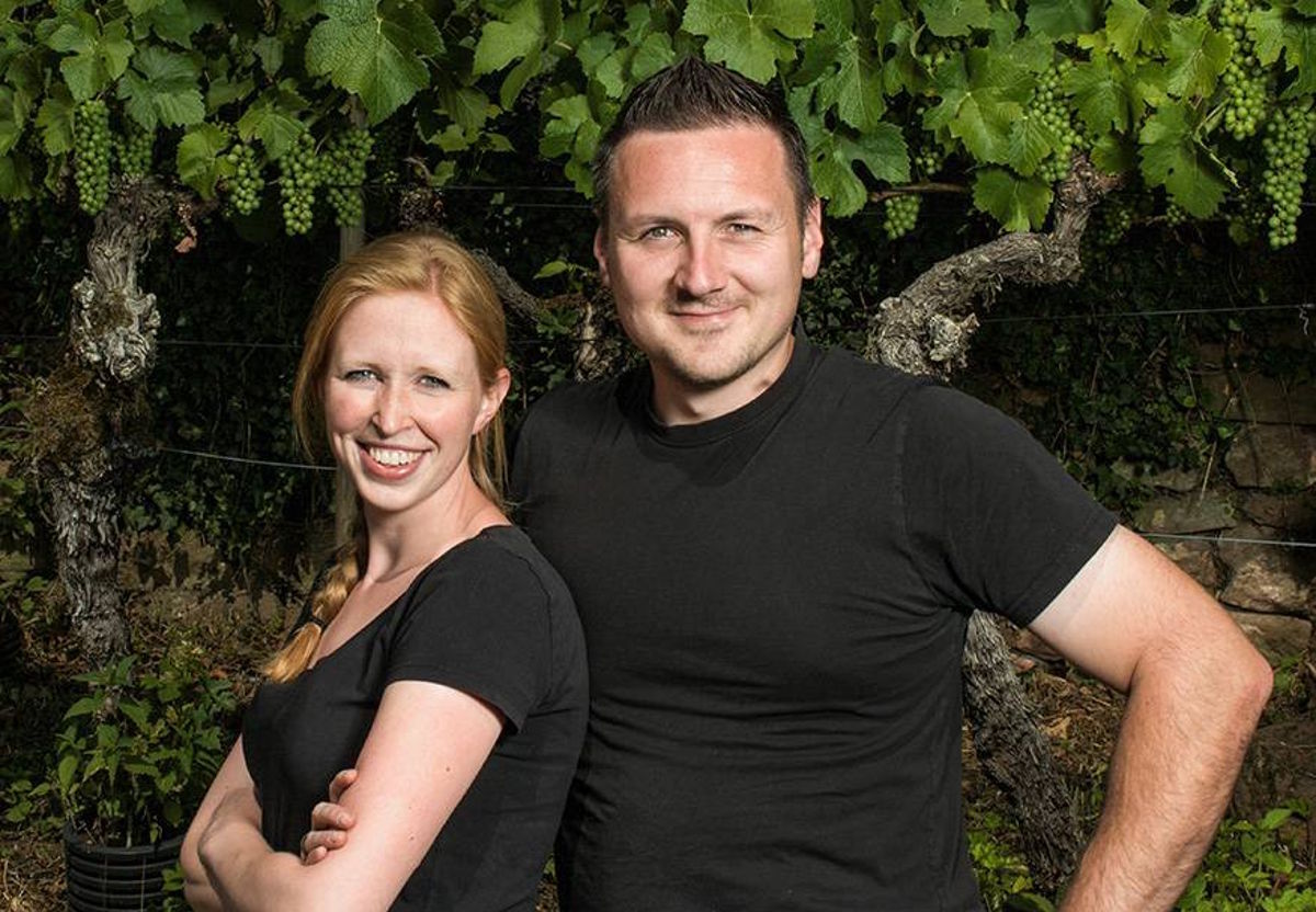 Lächelndes Paar in schwarzer Kleidung steht selbstbewusst vor einem Rebstock im Weinberg