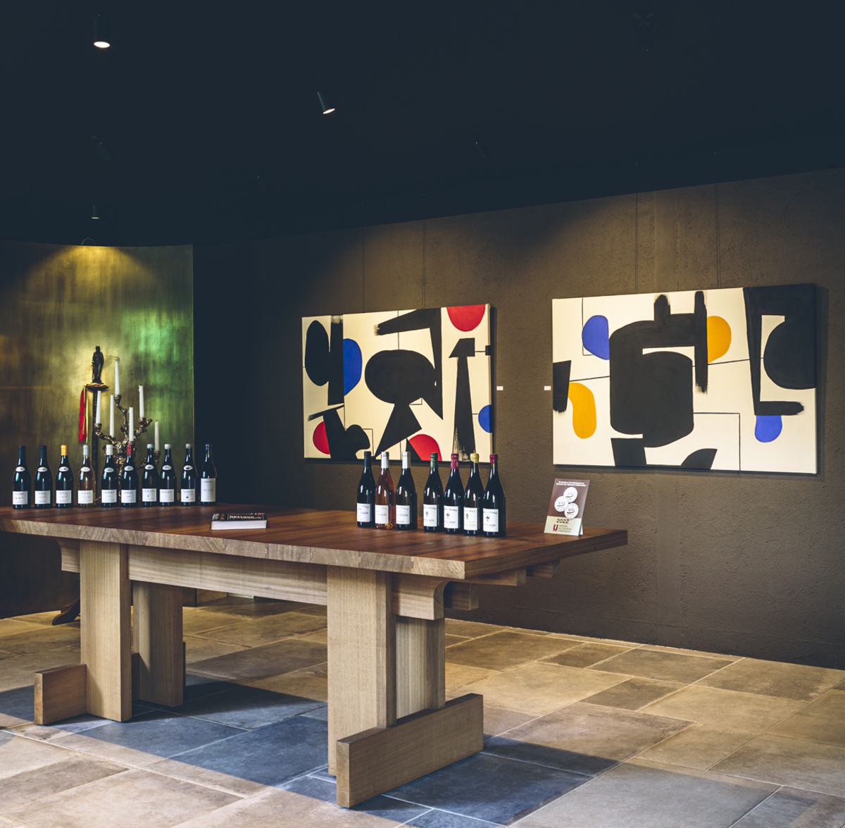 Holztisch mit Weinflaschen in modern gestalteter Vinothek, im Hintergrund abstrakte Kunstwerke an dunkler Wand.