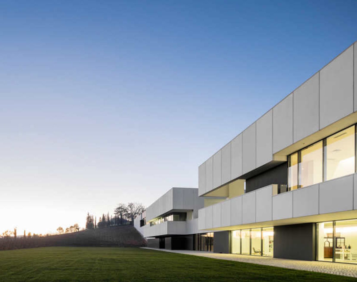 Modernes Weingutsgebäude mit klarer, geometrischer Architektur in heller Abendstimmung, davor eine gepflegte Rasenfläche.