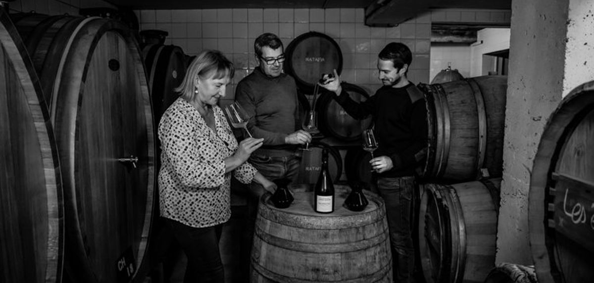 Drei Personen verkosten in einem Weinkeller zwischen großen Holzfässern, Gläser und Flasche auf einem Fass.