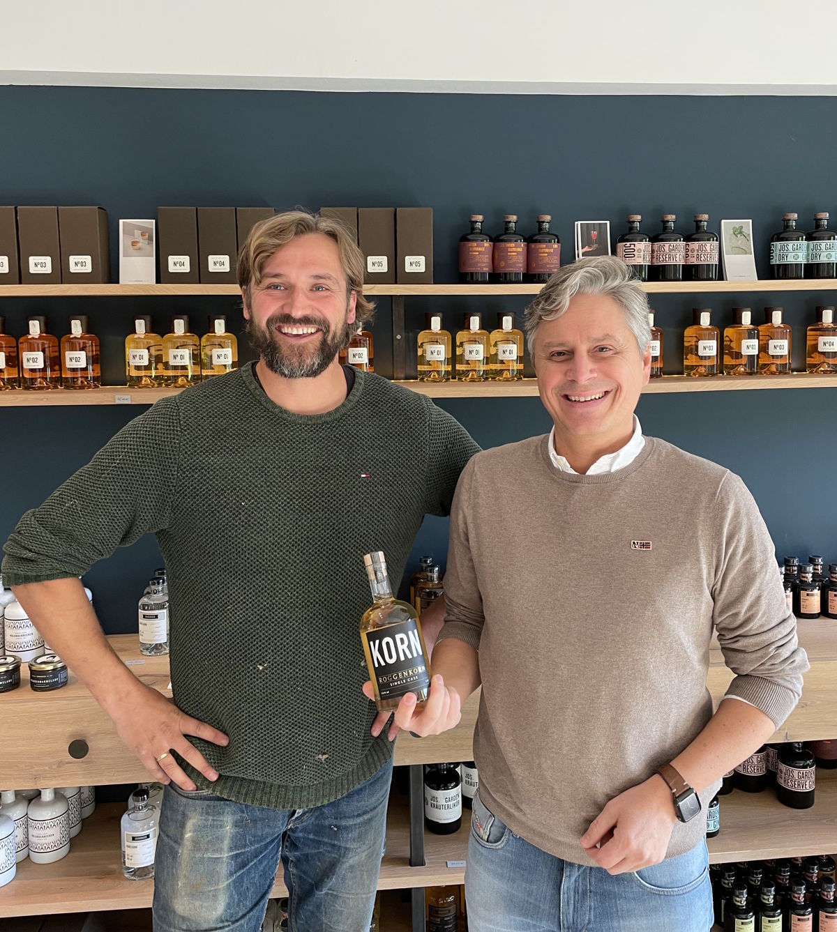 Zwei freundlich lächelnde Männer stehen in einem stilvollen Verkaufsraum und präsentieren stolz eine Flasche Kornbrand – im Hintergrund eine Auswahl hochwertiger Spirituosen auf Holzregalen