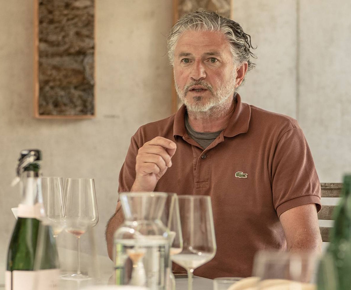 Mann mit grauem Haar und Bart sitzt an einem Tisch mit Gläsern, Flaschen und Karaffe, vor hellem Hintergrund.