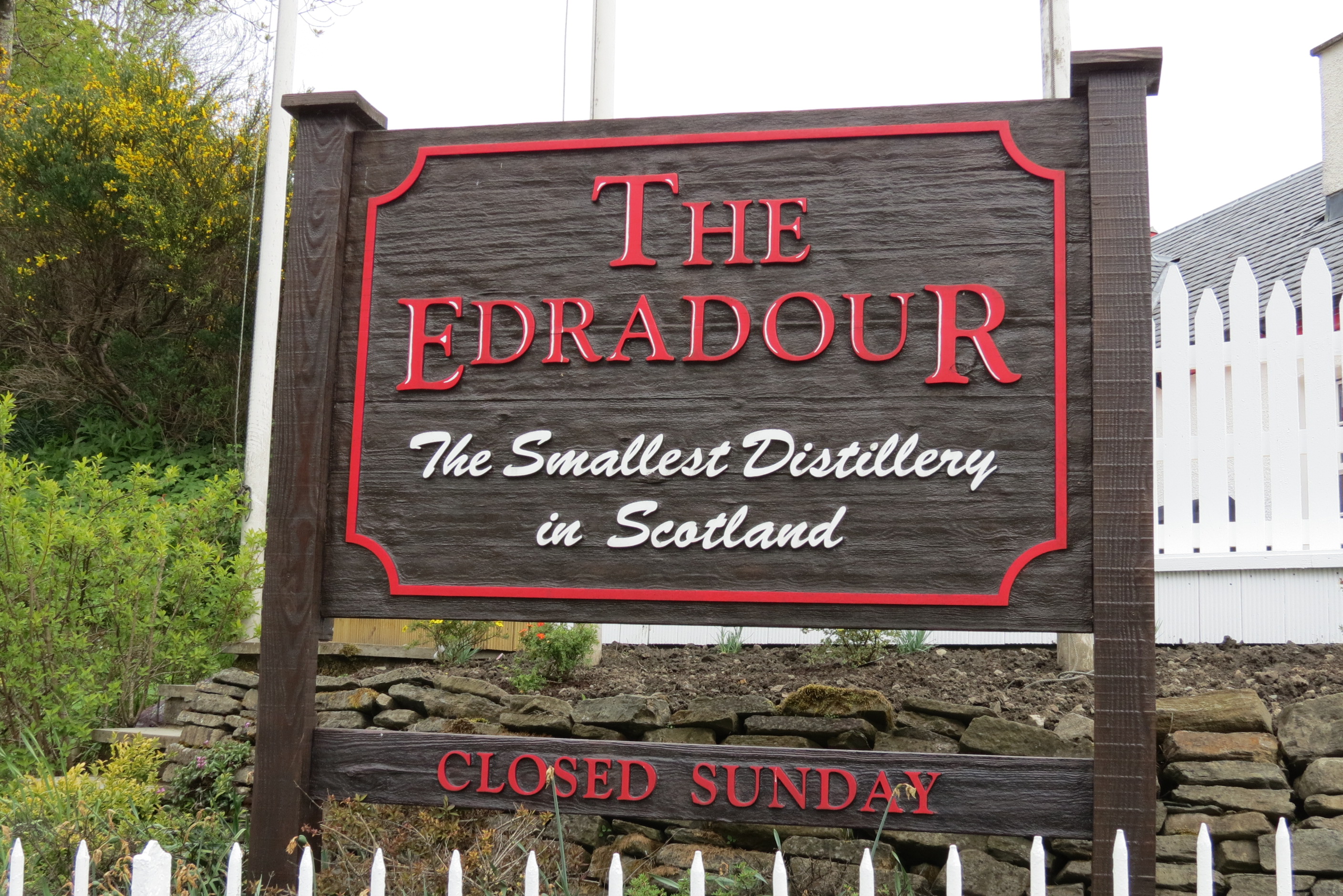 Holzschild mit roter Schrift „The Edradour – The Smallest Distillery in Scotland“ sowie Hinweis „Closed Sunday“, aufgestellt vor einem weißen Zaun und Grünanlage – markanter Eingangsbereich der bekannten schottischen Whisky-Destillerie