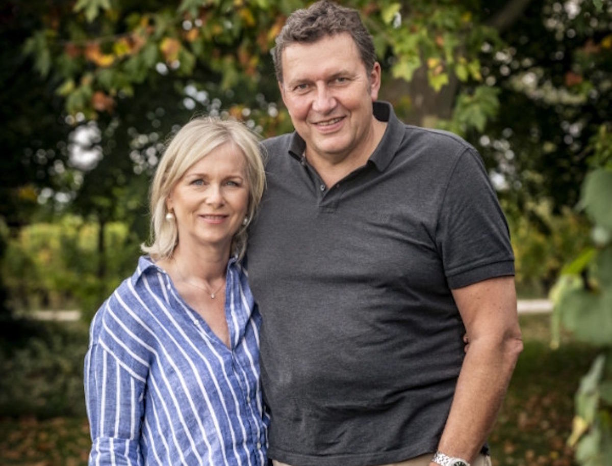 Winzerpaar steht lächelnd nebeneinander im Freien, im Hintergrund Bäume mit teils herbstlich gefärbtem Laub.