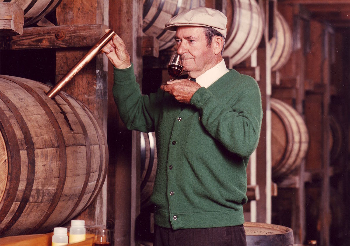 Ein älterer Mann entnimmt mit einer Pipette oder einem Weinheber eine Probe aus einem Holzfass und riecht daran – traditionelle Qualitätskontrolle während der Reifung von Wein oder Spirituosen im Fasslager