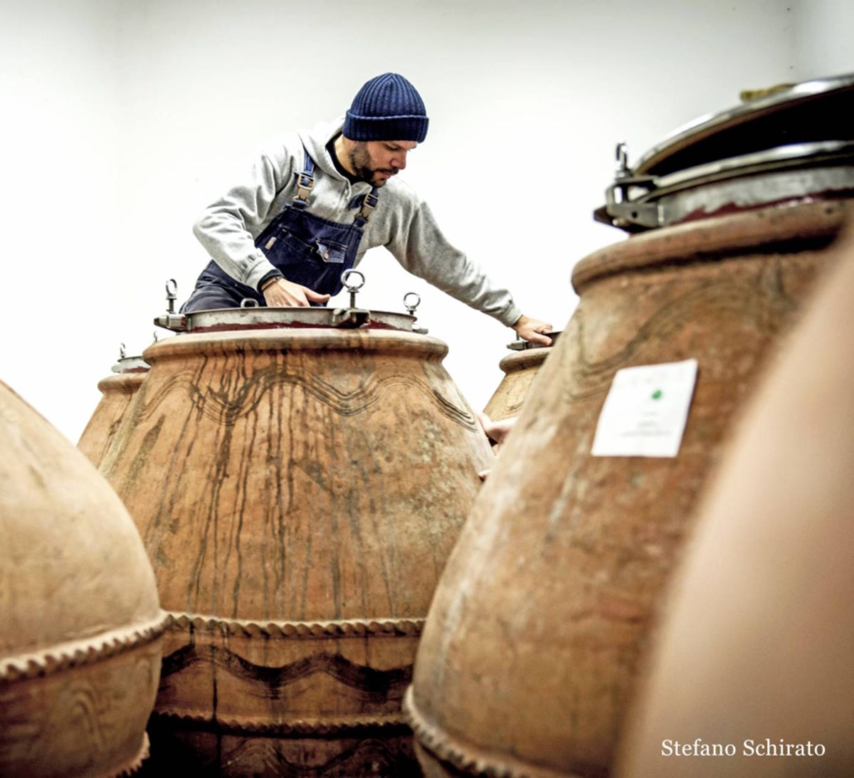 Winzer mit Mütze und Latzhose arbeitet an großen Tonamphoren mit Metallverschlüssen in einem modernen Kellerraum.